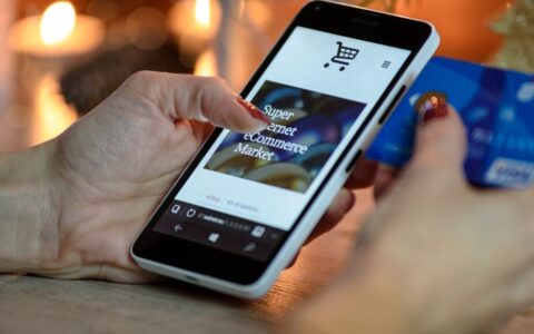 Woman using smartphone for online shopping with credit card in hand, festive background lighting.