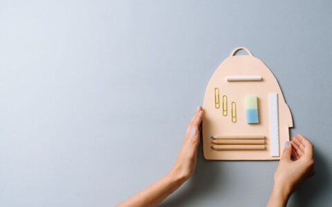 Flat lay of school supplies on a cutout in backpack shape, held by hands on a light surface.