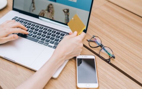 A person makes an online purchase using a credit card on a laptop, with a smartphone and glasses nearby.