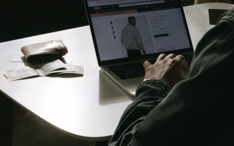 Adult man shopping for clothes online at home using a laptop, with a wallet on the table.