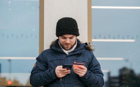 Positive man in warm clothes standing near building and entering credentials of credit card on mobile phone in daylight