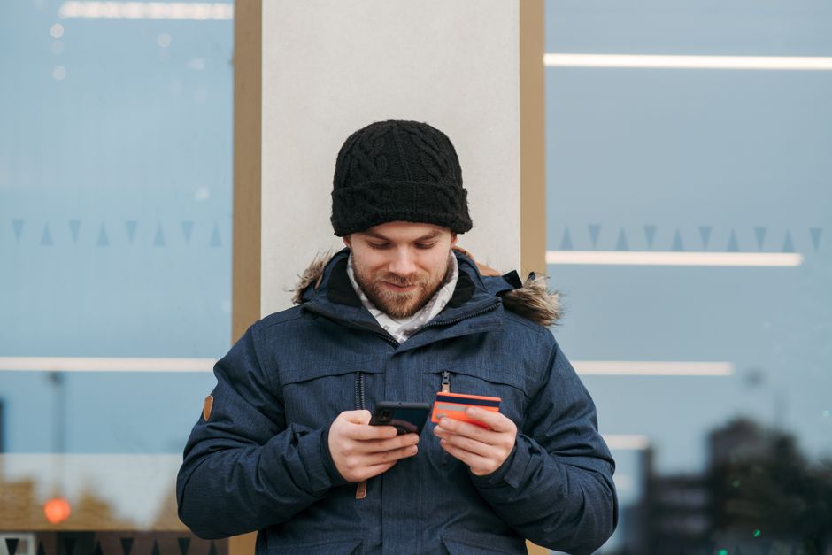 Positive man in warm clothes standing near building and entering credentials of credit card on mobile phone in daylight