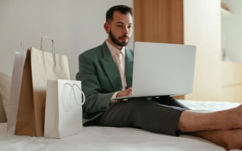 Adult man in teal blazer shopping online using laptop on bed with shopping bags.