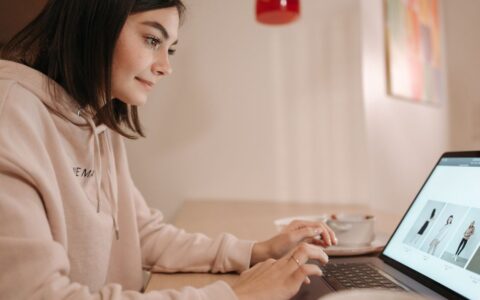 Young woman in pink hoodie browsing online fashion store on laptop at home. Calm and focused atmosphere.