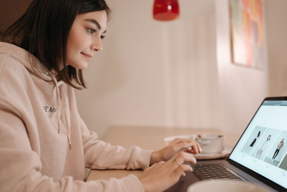 Young woman in pink hoodie browsing online fashion store on laptop at home. Calm and focused atmosphere.