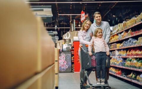 Family enjoying a shopping trip in a supermarket, with a child in a cart and parents smiling.