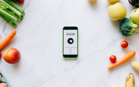 Mobile delivery app surrounded by fresh fruits and vegetables on a marble countertop.