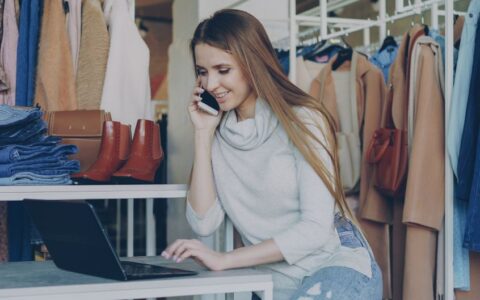 Smiling woman in a fashion boutique using a laptop and phone.