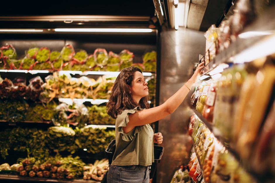 A woman selects fresh vegetables in a well-lit supermarket produce aisle.