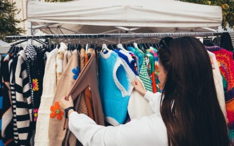A woman browsing colorful vintage sweaters at an outdoor market stall.