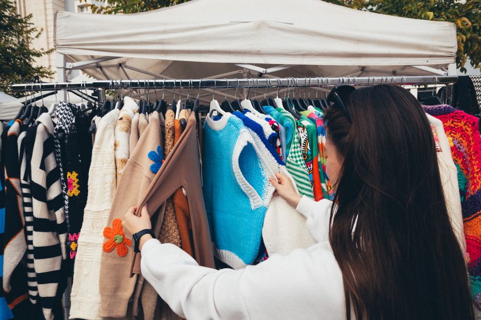 A woman browsing colorful vintage sweaters at an outdoor market stall.